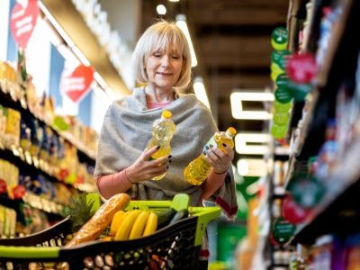 femme qui regarde deux bouteilles d'huile. elle est dans un rayon de supermarché derrière un caddie rempli de nourriture