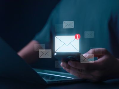 main tenant un téléphone au-dessus du clavier d'un ordinateur portable. Au dessus des enveloppes dont une avec un point d'exclamation