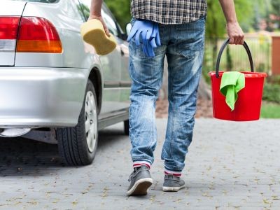 bas du buste, jambes d'un homme et bras avec dans les mains une éponge et un seau. Dans la poche de son pantalon des gants en plastique. Une voiture garée devant lui.