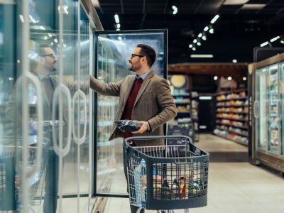homme devant un caddie tenant une bouteille et regardant dans un placard vitré ouvert dans un rayon de supermarché