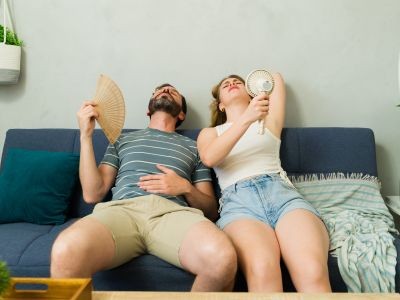 Couple assis sur un canapé essayant de se rafraîchir avec un éventail et un ventilateur portable pendant une forte chaleur.