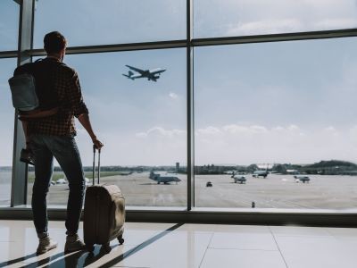 homme tenant une valise dans un hall d'aéroport regardant des avions dont un qui décolle