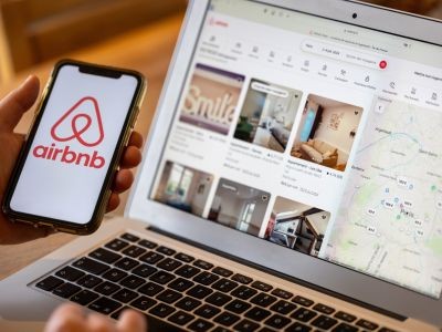 main tenant un smarphone avec le logo airbnb sur l'écran. Orinateur portable ouver posé sur une table avec sur l'écran des annonces